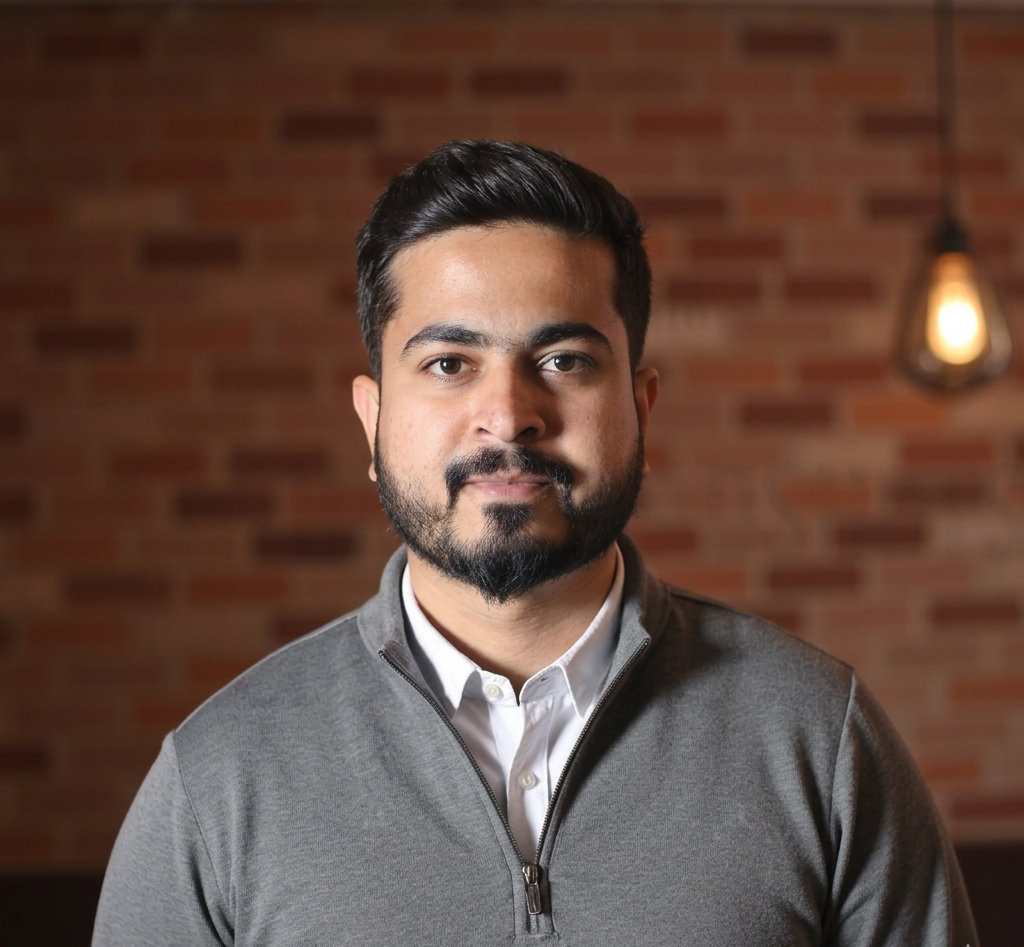 Portrait of a man with dark hair and a trimmed beard, wearing a white shirt and gray zip sweater, standing against a brick wall.